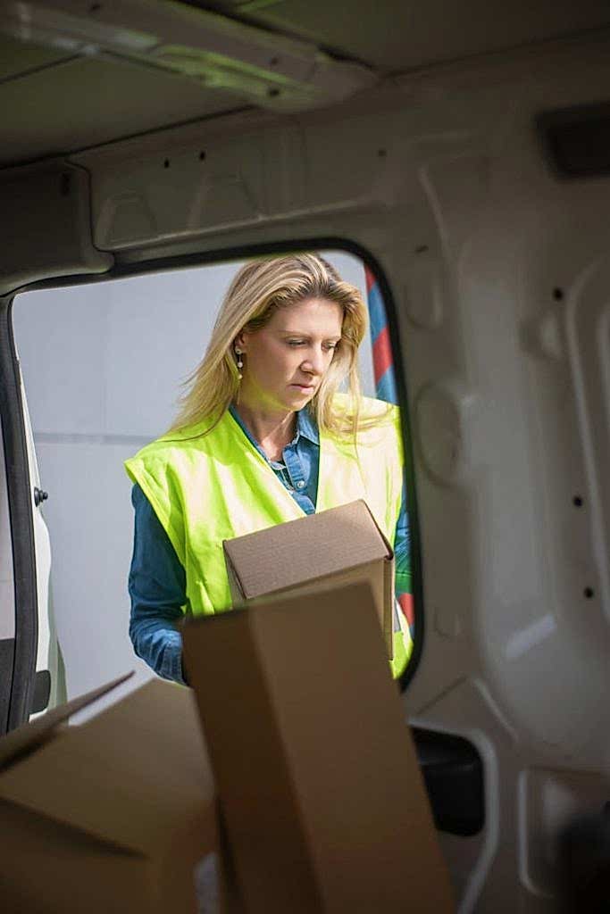 woman-in-blue-long-sleeve-shirt-and-green-vest-standing-beside-white-vehicle-7843953 Woman in high-visibility vest loading packages inside a delivery van.